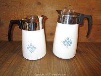 Two white Corning Ware coffee pots side by side showing front view with blue floral pattern, black handles, and stainless steel tops.