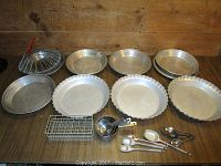 Photo showing 10 aluminum pie plates arranged on a wooden surface including three wavy edged plates and seven plain plates; metal folding wire strainer, measuring cups, spoons, and a metal rack.