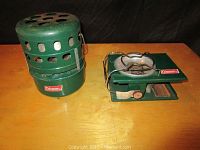 Photo showing the green Coleman model 510A heater and the small green Coleman camping stove on a wooden surface against a black backdrop.