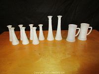 Nine milk glass vases, seven approximately 6 inches tall and two approximately 9 inches tall, plus two large mugs on a wooden surface with dark background.
