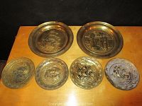 All six vintage brass hanging plates arranged on a surface, showing variety of embossed designs including ships, a peacock, and figures.