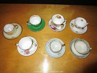 Top-down photo of 7 tea cups with matching saucers arranged in 2 rows, showing different floral and gold-trimmed patterns