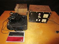 Photo showing Kodak Carousel slide projector alongside two wooden slide trays and original user manual on wooden table.