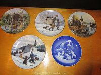 Top-down view showing all five decorative hanging plates on a wooden surface. Four plates feature Keirstead style winter and rural scenes; fifth plate is blue with dog artwork.