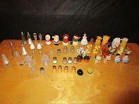 Photo showing a variety of salt and pepper shakers arranged on a wooden surface including glass and ceramic novelty designs.