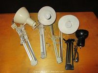 Four extendable lamps with visible cords and clamp bases placed on a wooden surface. Two lamps are off-white, one is white, and one is black.