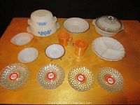 Top view of all Fire King dishes including casseroles, candy dishes, covered dish, orange mugs, divided serving bowl and sixteen crystal plates spread out on a wooden table.