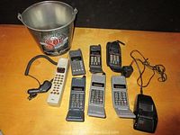 Photo of seven vintage cell phones and two chargers arranged next to a branded Sol beer metal bucket on a wooden surface.