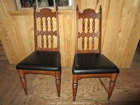 Front view of two wooden chairs with carved backs and black upholstered seats, placed against a wooden wall.