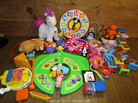 Assorted girl’s toys on a wooden surface including plush unicorn, My Little Pony figures, Mickey Mouse learning clock, small animal figures, musical instruments, and toy blocks.