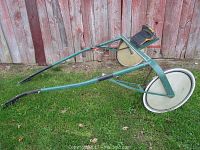 Side view of green metal harness racing sulky showing frame, wheels, and seat.