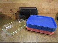 Two rectangular transparent glass Pyrex baking dishes positioned at front left with stacked colorful plastic vintage restaurant trays to the right and behind them on a wooden surface.