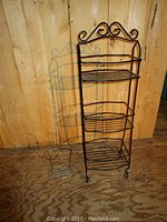 Photo of two metal stands against wood panel background, showing taller black metal bathroom storage stand and smaller silver-tone metal basket stand on floor.