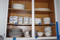 Photo showing white ceramic dishware with blue line and brushstroke patterns including stacked plates, bowls, and multiple tea cups arranged inside a wooden cabinet.