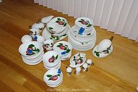 Overview photo of entire dish set showing various plates, bowls, cups, and serving pieces with hand painted red apple design and green leaves on white ceramic.
