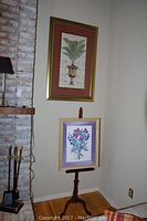 Two framed botanical prints shown on wall and easel, one large palm print in gold frame on wall and smaller floral print in light wood frame on easel.