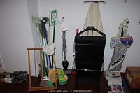 Full view of vintage drying rack, assortment of cleaning tools (mops, brooms, dusters), Shark steam mop manual attached to unit, Dirt Devil Swift Stick vacuum, Oreck vacuum cleaner, pack of cleaning cloth refills, and shoe collection on floor.