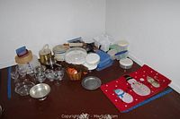 Photo showing assorted kitchen items on the floor against a white wall, includes glassware, metal platter, plastic containers, bowls, cream canisters, woven basket, and a red holiday placemat with snowman design.