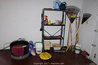 Photo showing garden rakes, garden supplies like soil bags, watering can, a black metal shelf with various hand tools and accessories, and jumper cables on the floor.