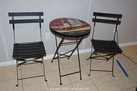 Set including one round folding bistro table with a colorful top and two matching folding metal chairs with black slats.