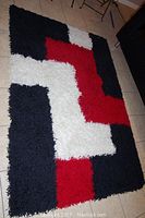 Full view of red, white, and black geometric shag area rug laid flat on tiled floor, showing overall shape and pattern.