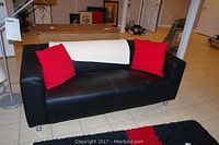 Full view of black leather loveseat with two red pillows and an ivory throw on tiled floor.