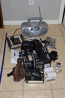 Wide view of the lot showing the Philips CD sound machine, Canon EOS Rebel X with lens, battery charger boxes, camera bags, and other accessories spread on a tile floor.