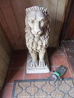 Concrete lion statue in corner on tiled floor next to doormat, also showing small metal cricket figurine on floor.