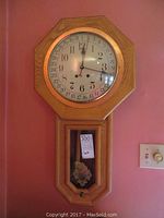 Front view of reproduction oak school wall clock with round white face, black numerals, copper ring, and pendulum visible behind glass panel.