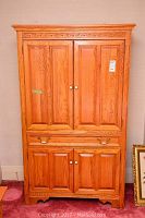 Front view of the oak armoire closed, showing four paneled doors, one drawer underneath the top doors, brass handles on the drawer, and carved detail along the top trim.