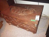 Front and top view of wooden chest with carved floral designs on lid and front. Measurements tag visible.