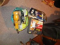 Complete lot overview showing the orange handheld sander, black tool case, green bag and hacksaw on a carpet .