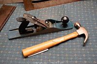 Photo showing a wood handle claw hammer, a blue metal hand planer, and a large metal square saw blade all resting on a patterned blue cloth.