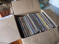 Open cardboard box filled with numerous vinyl records organized vertically.