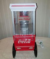Front view of Coca Cola branded popcorn maker showing transparent compartment with text 'Enjoy That Refreshing New Feeling!!' and red metal lower section with white Coca Cola logo, black wheels, and white top with red stripes.