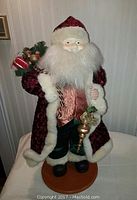 Front view of the Santa Claus figurine showing the velvet burgundy coat, white faux fur trim, fluffy beard, metallic vest, and the gift bag with greenery and small wrapped presents