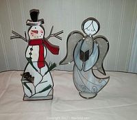 Stained glass snowman and angel candle holders displayed side by side on white surface against paneled wall.