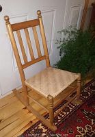 Youth size wooden rocking chair made of maple with vertical slat back and new woven splint seat. Natural wood finish with turned finials on the back posts. Chair placed on wooden floor next to a door and rug.