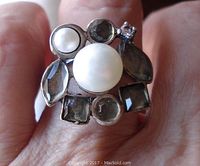 Close-up front view of sterling silver ring worn on finger showing two cultured pearls—a larger 10 mm and smaller 5 mm—along with one tiny clear stone and six different shaped light green bezel-set stones.