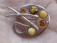 Front view of vintage sterling silver painter palette brooch held in hand showing five amber stones of varying colors and two brushed crossing the palette.