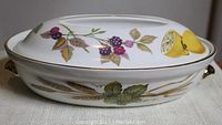 Side view of oval serving dish with lid showing fruit and wheat decoration and gold trim.