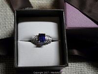 Front view of 10K white gold ring with synthetic blue sapphire in white leatherette ring box and outer purple box with brown ribbon.
