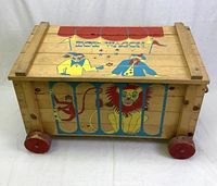 Front view of a rectangular wooden toy chest wagon with painted clowns, circus tent roof, and text 'Toy Wagon' on the lid. Side painted with monkey, lion behind blue bars. Red wooden wheels underneath.