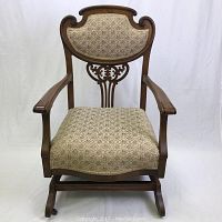 Front view of antique wooden rocking chair with upholstered seat and backrest, showing carved wood detail and wooden armrests.