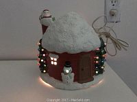 Ceramic Christmas cottage glowing with light-up trees and snowman illuminated. Power cord visible at back.