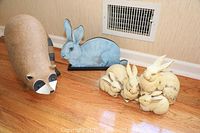 Photo showing three yard ornaments: heavy raccoon on the left, blue wooden bunny with stand in the center back, and cream-colored bunnies grouped on the right.