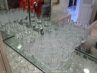 13 clear old fashioned crystal glasses arranged on a glass shelf reflecting in a mirror. Glasses show diamond cut and vertical cut faceted patterns.