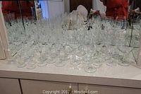 Multiple rows of clear stemware glasses with wide bowls and tall stems arranged on a white surface in front of a mirror.