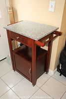 Granite top kitchen cart showing front and side with drawer, shelf and cabinet door, wooden frame with metal knobs and side handles, caster wheels underneath.