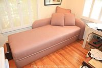 Full view of the chaise, upholstered in textured dusty rose fabric, with three matching pillows arranged at the backrest, positioned near a window and a desk with papers.